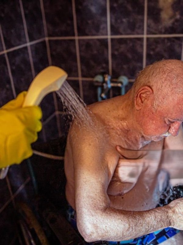 In the bathroom, caring male nurse in protective workwear helps to a senior male patient in wheelchair to bath himself