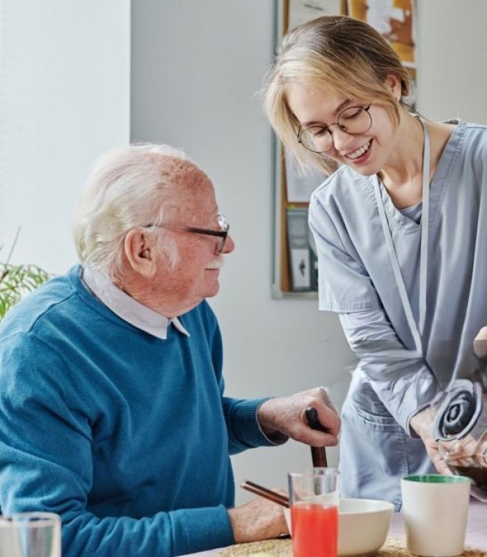 Young caregiver pouring coffee from teapot for senior man while he having breakfast at table