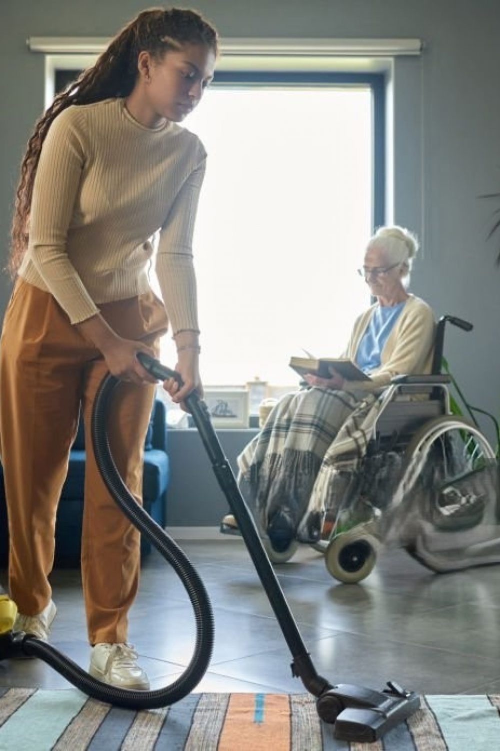 Cute teenage girl in casualwear cleaning floor of living room with vacuum cleaner against her grandmother with disability sitting in wheelchair