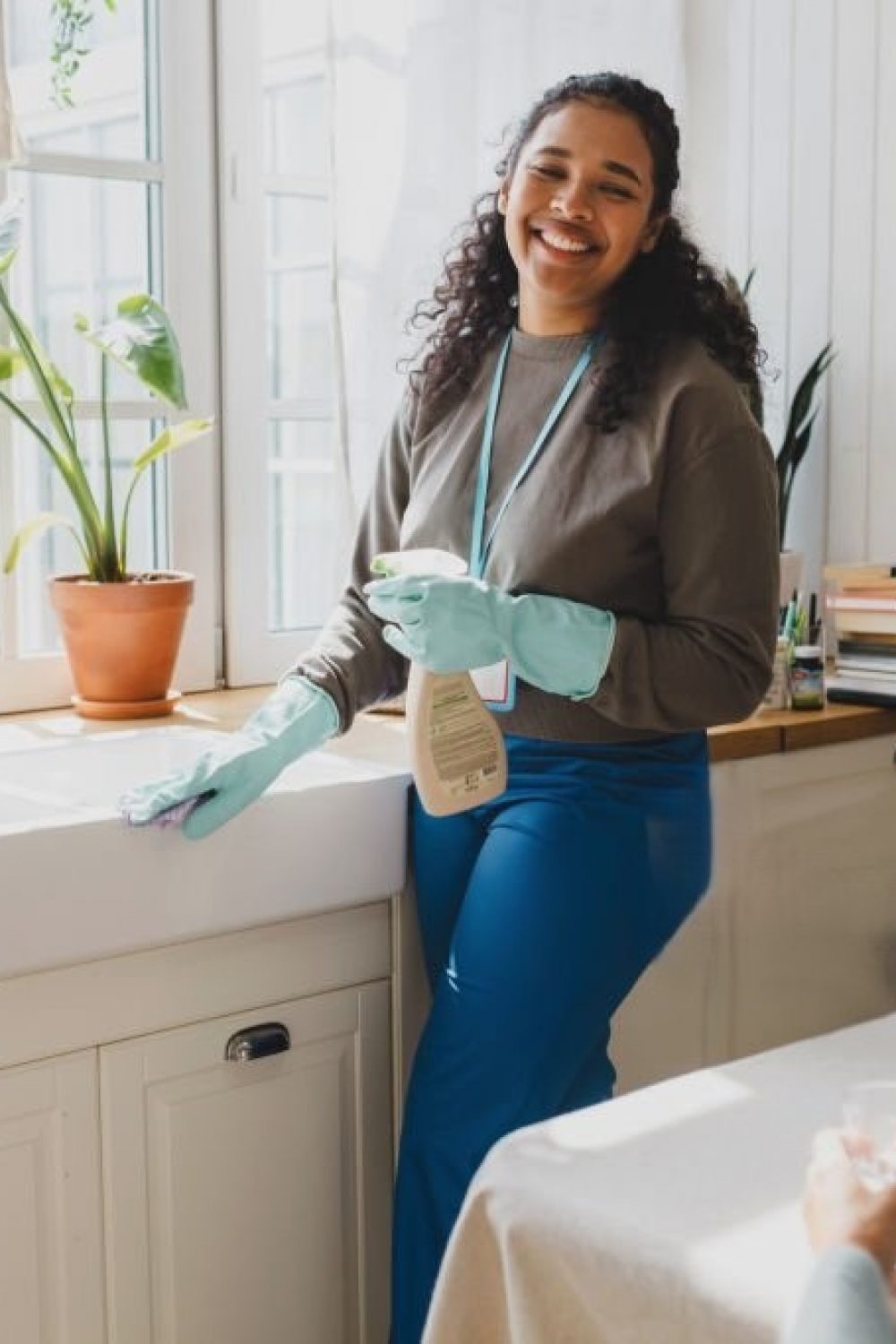 Selective focus on happy african american volunteer helping senior lady to clean house, standing in kitchen in raisin gloves washing sink with detergent, talking to elderly female sitting at table